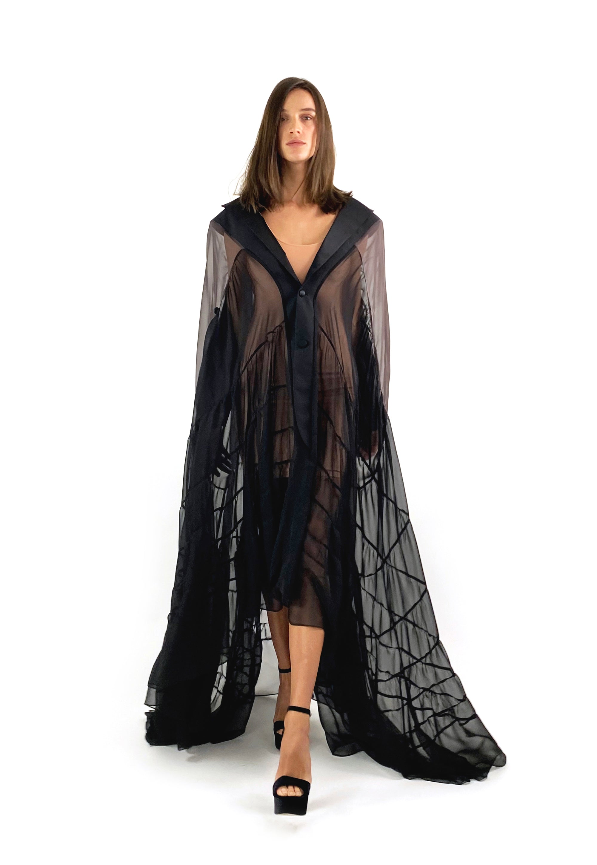 A model showcases a black sheer cape paired with elegant black heels, exuding sophistication and style.