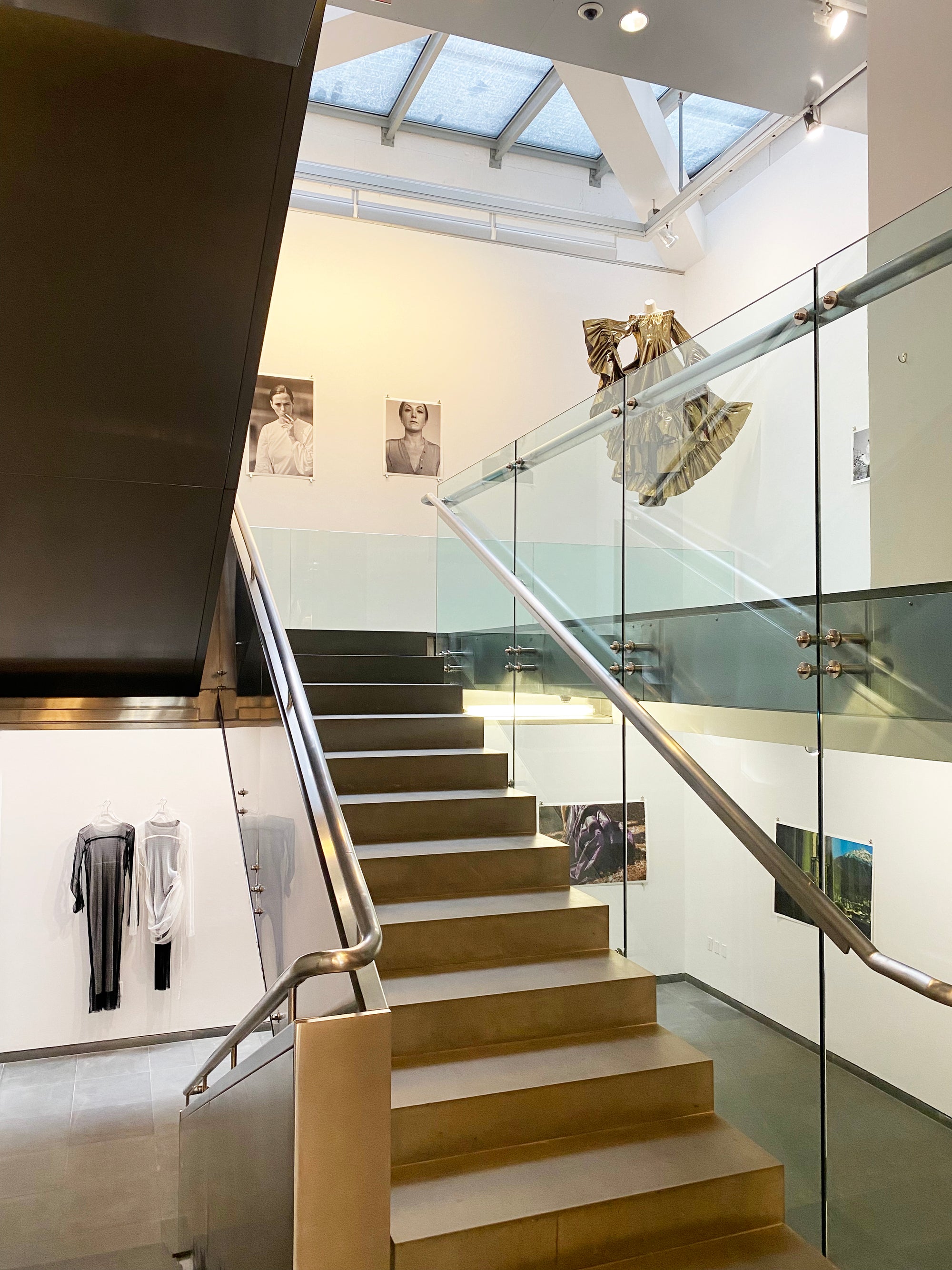 A modern stairway featuring a glass railing alongside a wall adorned with various framed pictures and dresses.