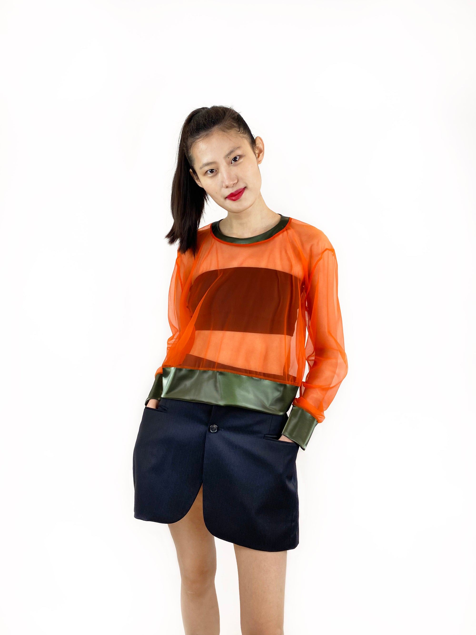 A stylish woman in a sheer orange top with shiny green cuffs and a black skirt poses elegantly, highlighting her fashion sense and confidence.