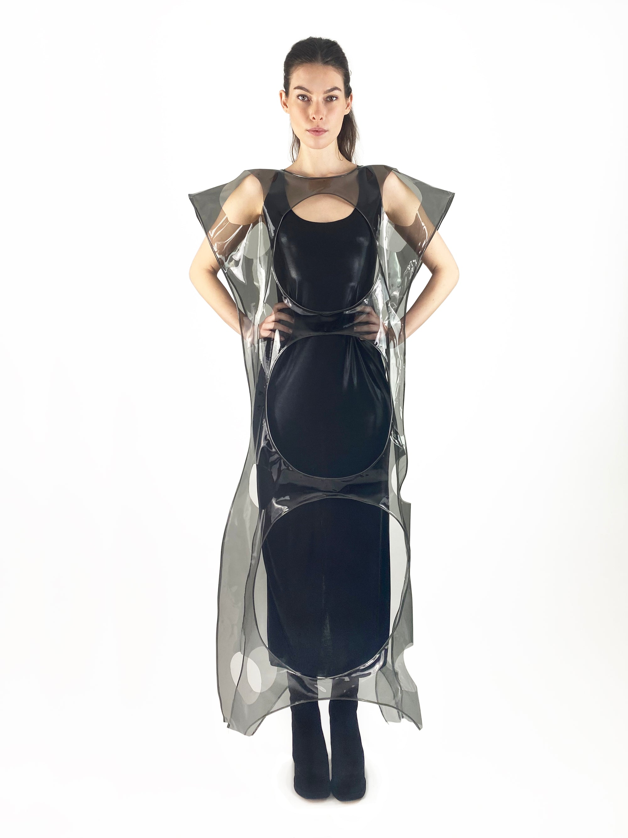 A woman elegantly dressed in a black dress and a plastic cape with circular cut-outs, exuding sophistication and mystery.