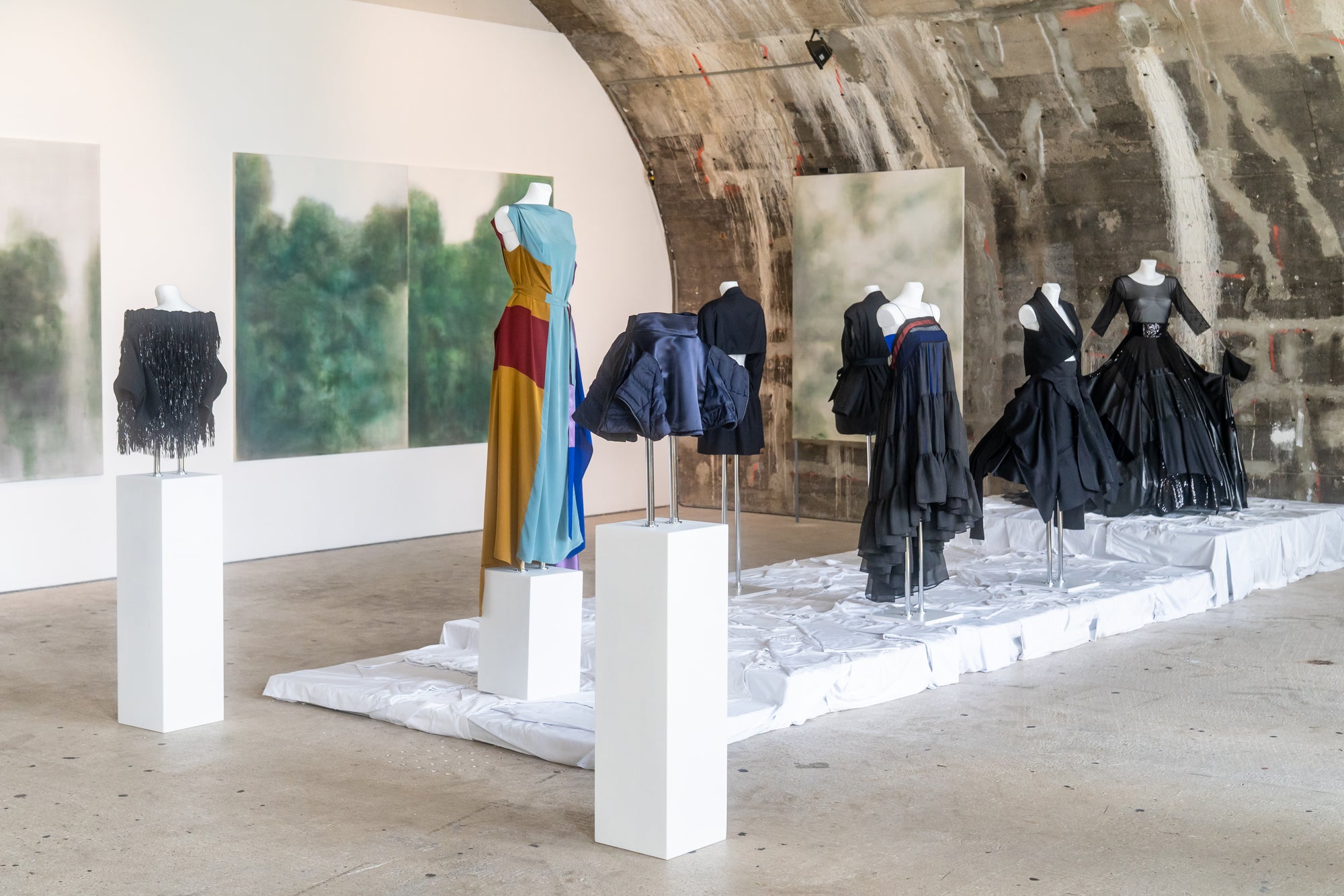 Various stylish dresses draped on mannequins on white pedestals in an art gallery environment. 