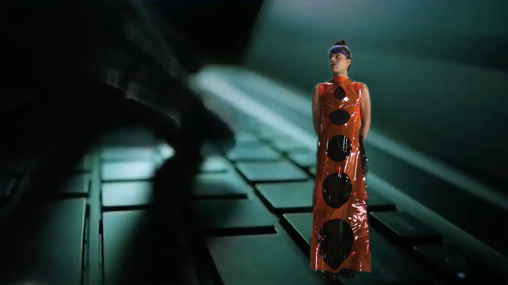 A woman in an orange plastic dress stands atop a laptop, symbolizing the intersection of technology and fashion.