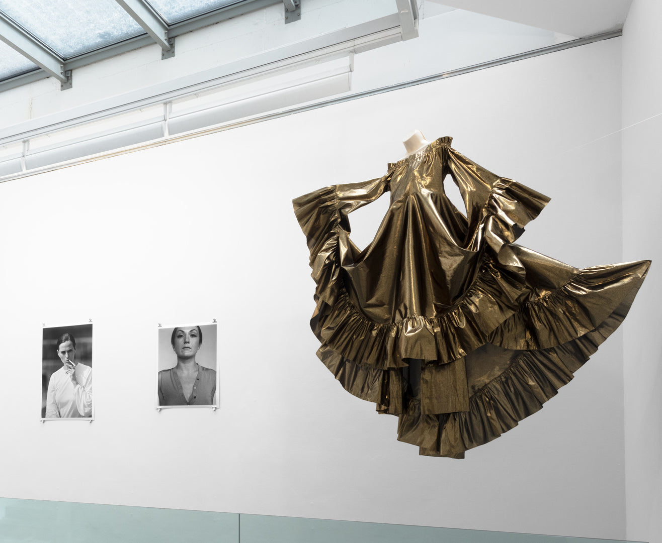  A large gold dress elegantly suspended from the ceiling next to two photographs of portraits, showcasing its shimmering fabric and intricate design.